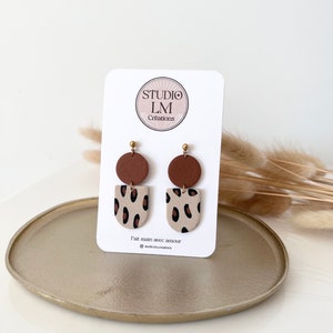 Peut inclure: Une paire de boucles d'oreilles pendantes marron et beige avec un motif léopard. Les boucles d'oreilles sont faites à la main et présentent un cercle marron en haut et une demi-lune beige à imprimé léopard en bas. Les boucles d'oreilles sont sur une carte blanche avec le texte "Studio LM Creations" et "Fait main avec amour" en français.