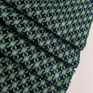 May include: Close-up of folded fabric with a repeating chain-link pattern. The fabric has a navy blue background with interlocking circles in shades of green and teal. The folds show the texture and depth of the material, creating a visually interesting display.
