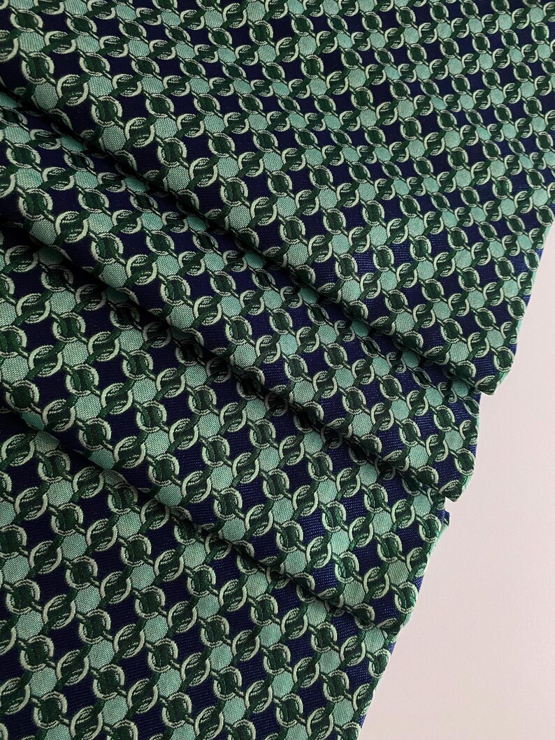 May include: Close-up of folded fabric with a repeating chain-link pattern. The fabric has a navy blue background with interlocking circles in shades of green and teal. The folds show the texture and depth of the material, creating a visually interesting display.
