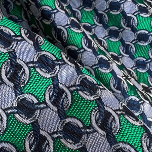 May include: Close-up of a fabric with a repeating chain pattern. The design features interlocking chain links in shades of blue, green, and navy. The fabric has a textured appearance, with a woven or textured surface.