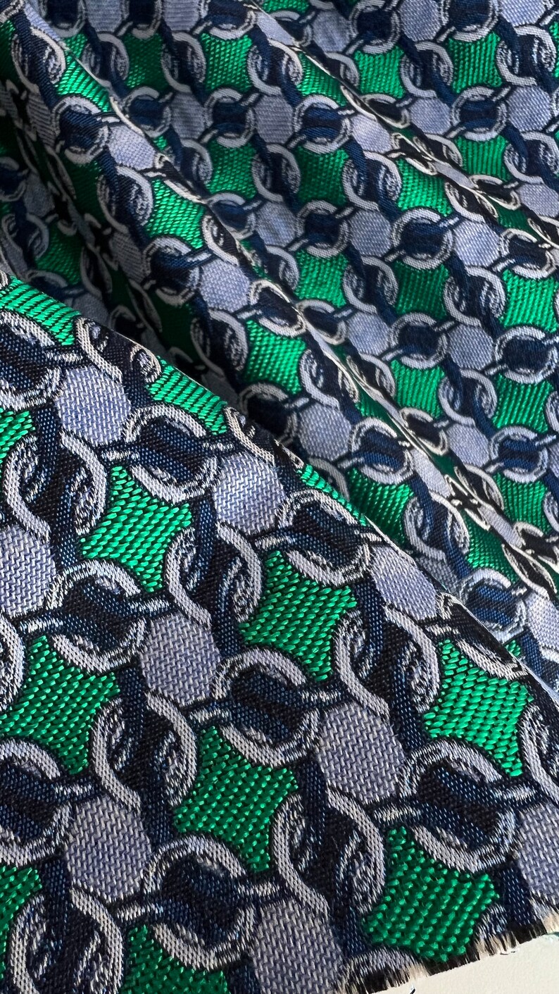 May include: Close-up of a fabric with a repeating chain pattern. The design features interlocking chain links in shades of blue, green, and navy. The fabric has a textured appearance, with a woven or textured surface.