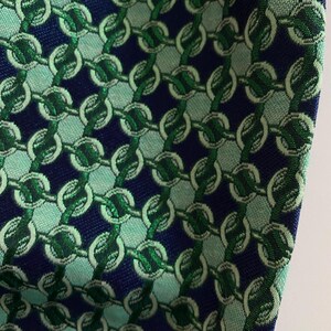 May include: Close-up of a patterned fabric with a repeating chain design. The fabric features a navy blue background with vertical rows of interlocking chains in shades of green and light teal. The texture of the fabric is visible.