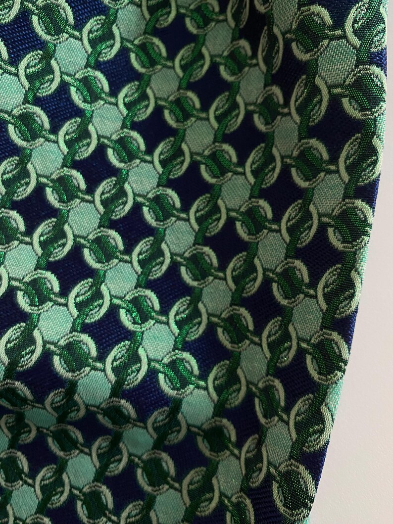 May include: Close-up of a patterned fabric with a repeating chain design. The fabric features a navy blue background with vertical rows of interlocking chains in shades of green and light teal. The texture of the fabric is visible.