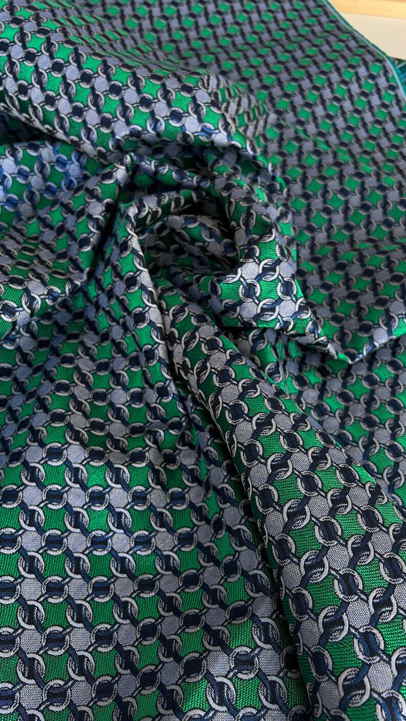May include: A close-up shot of a fabric with a chain-link design. The fabric is a mix of green, grey, and navy blue. The material is likely intended for clothing or home furnishings.