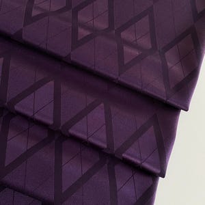 May include: Close-up of folded fabric in a deep purple hue, featuring a repeating diamond pattern in a darker shade. The fabric has a subtle sheen and a textured appearance, creating a visually interesting design.
