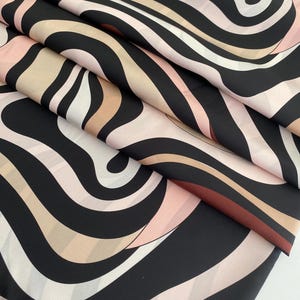 Könnte beinhalten: Ein Stoffstück mit einem sich wiederholenden abstrakten Muster aus fließenden, wellenförmigen Linien in den Farben Schwarz, zartem Rosa, Beige und Dunkelbraun. Das Material ist in Schichten gefaltet und bildet eine Reihe diagonaler Falten.