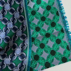 May include: Close-up of two fabric swatches with detailed patterns. One has a chain link design in green, blue, and silver. The other shows a floral pattern in green and dark green on a grey background. Both have fringed edges.