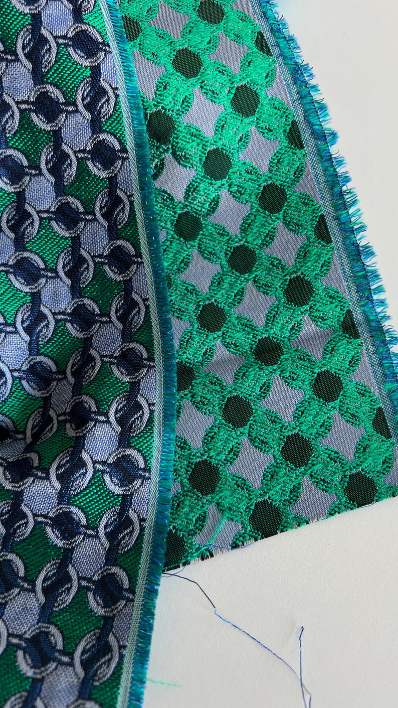 May include: Close-up of two fabric swatches with detailed patterns. One has a chain link design in green, blue, and silver. The other shows a floral pattern in green and dark green on a grey background. Both have fringed edges.