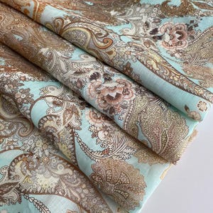 May include: Folded fabric with a detailed paisley pattern. The fabric has a light blue base, overlaid with brown, beige, and pink floral and paisley motifs. The folds create shadows, emphasizing the intricate design and colour variations.