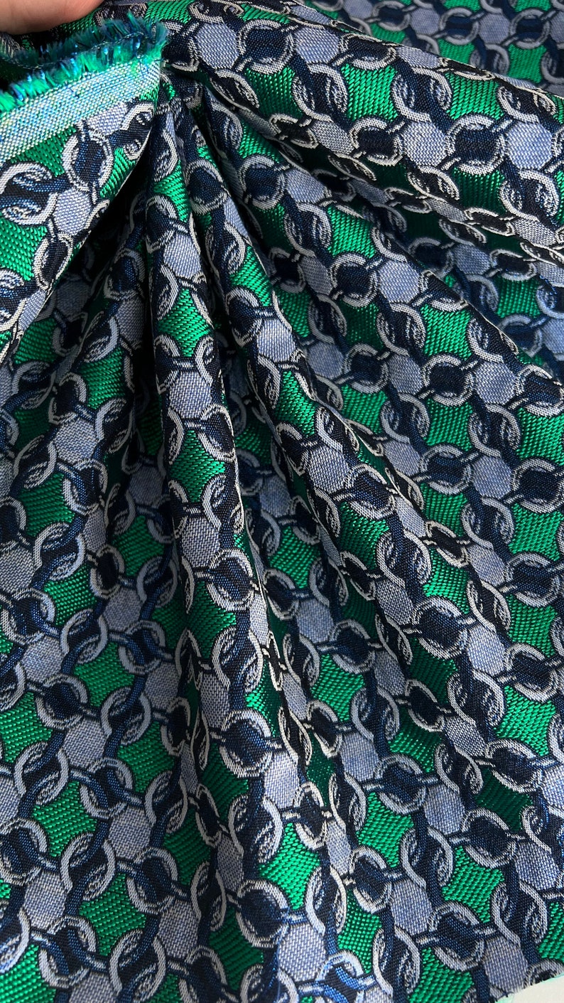May include: Close-up of a fabric with a chain link pattern. The fabric features a repeating design of interlocking circles in shades of green, blue, and grey. The fabric is folded, showing texture and depth.