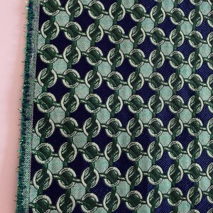 May include: Close-up of a fabric sample with a repeating chain-link pattern. The design features interlocking circles in shades of green and white against a dark blue background. The edge of the fabric has a green fringe.