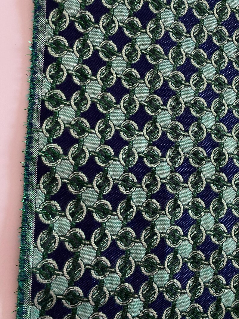 May include: Close-up of a fabric sample with a repeating chain-link pattern. The design features interlocking circles in shades of green and white against a dark blue background. The edge of the fabric has a green fringe.