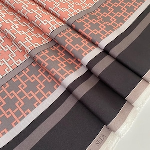 May include: Folded fabric with a geometric pattern in coral, gray, and white. The fabric has a border of gray and black stripes. The word "SETA" is visible on the edge of the fabric.