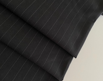 May include: Close-up of folded black fabric with thin, light-colored pinstripes. The fabric is layered, revealing the texture and pattern. The material appears to be a tailored suiting fabric, suitable for garments like trousers or jackets.