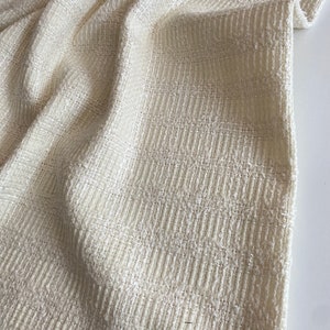 May include: Close-up of a white textured fabric with a subtle sheen. The fabric has a woven pattern and a slightly rough texture.