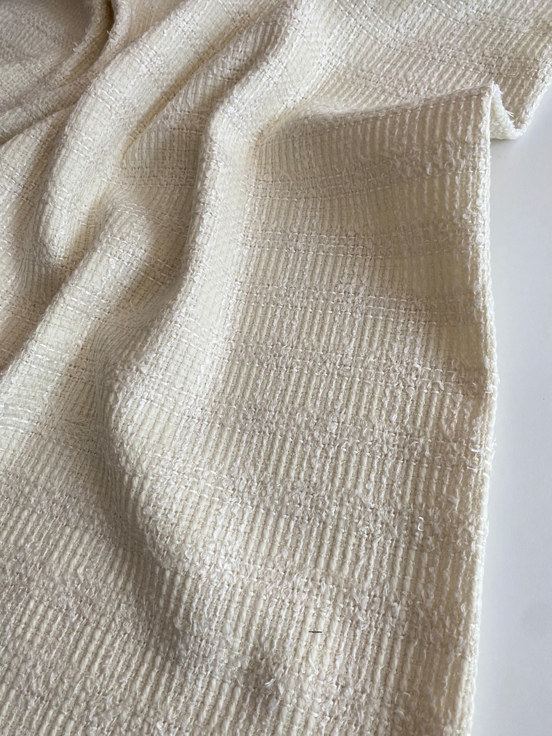 May include: Close-up of a white textured fabric with a subtle sheen. The fabric has a woven pattern and a slightly rough texture.