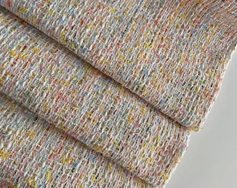 Puede incluir: Primer plano de una tela tejida con una superficie texturizada. La tela es principalmente blanca con motas coloridas de amarillo, rosa, azul y naranja. El material está doblado sobre sí mismo, mostrando las capas del textil.
