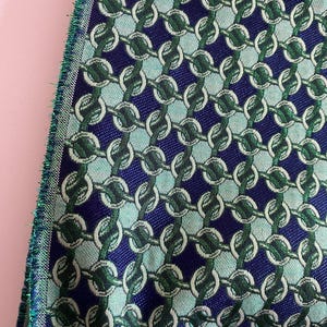 May include: Close-up of a woven fabric with a repeating chain link pattern. The fabric features a navy blue background with light blue and green chain links. The edge of the fabric has a green fringe.