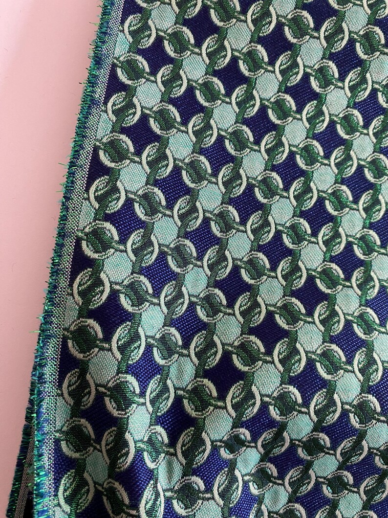 May include: Close-up of a woven fabric with a repeating chain link pattern. The fabric features a navy blue background with light blue and green chain links. The edge of the fabric has a green fringe.
