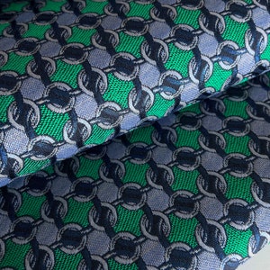 May include: Close-up of folded fabric with a repeating chain link pattern. The design features green, navy blue, and light blue colours. The fabric has a textured appearance, suggesting a woven material.