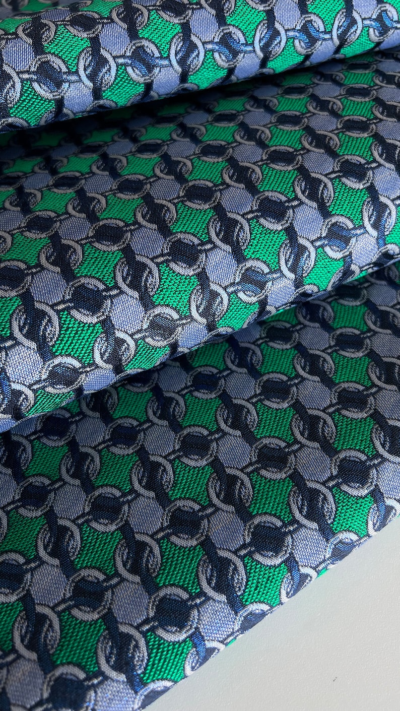 May include: Close-up of folded fabric with a repeating chain link pattern. The design features green, navy blue, and light blue colours. The fabric has a textured appearance, suggesting a woven material.