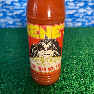May include: A bottle of red hot sauce with a label featuring the word "GENE" in yellow letters. The label also displays a black and white illustration of a face and the text "BITTER THAN HELL SAUCE".