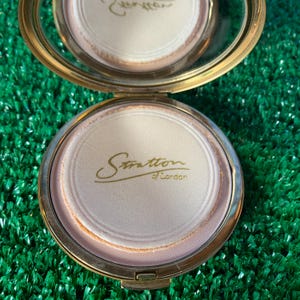 Op de afbeelding: Een gouden compact met een wit interieur en de tekst "Stratton of London" in goud gedrukt.