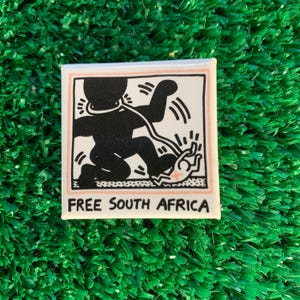 Puede incluir: Pin cuadrado con un gr&aacute;fico en blanco y negro de una persona con una cuerda alrededor del cuello, extendiendo la mano. El texto "FREE SOUTH AFRICA" est&aacute; debajo. El pin tiene un fondo blanco con un borde rosa.