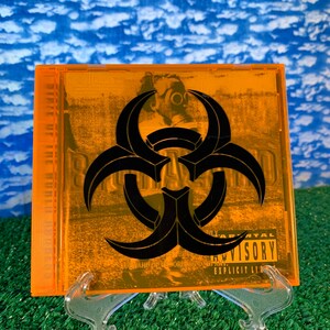 Op de afbeelding: Oranje CD-hoes met een zwart biohazard-symbool op de voorkant. De hoes heeft een Parental Advisory-sticker in de rechterbenedenhoek. De tekst op de sticker luidt "Parental Advisory Explicit Lyrics".