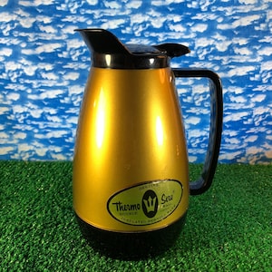 Op de afbeelding: Een gouden, dubbelwandige, geïsoleerde serveerkan met een zwarte deksel en handvat. De kan heeft een label met de tekst "Genuine Thermo Serv Double Wall Insulated Serving Ware".