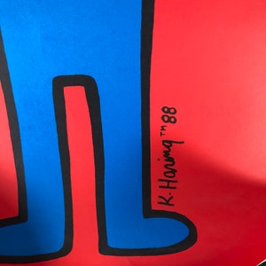 May include: A blue figure with black outlines stands on a red background. The figure's legs are visible and the signature "K. Haring TM '88" is written in black on the red background.
