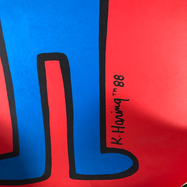 May include: A blue figure with black outlines stands on a red background. The figure's legs are visible and the signature "K. Haring TM '88" is written in black on the red background.