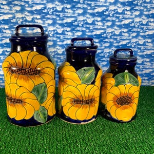 Könnte beinhalten: Set aus drei Keramikbehältern mit Deckeln. Die Behälter sind marineblau mit gelben Sonnenblumenmotiven und grünen Blättern. Jeder Behälter hat einen Griff am Deckel. Die Behälter sind in verschiedenen Größen erhältlich.