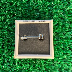 Puede incluir: Pin cuadrado con fondo negro y borde blanco. El pin tiene un cierre de imperdible plateado en la parte trasera. El texto "&copy; 1985 KEITH HARING" est&aacute; impreso en blanco en la parte superior. El pin est&aacute; sobre una superficie verde texturizada.