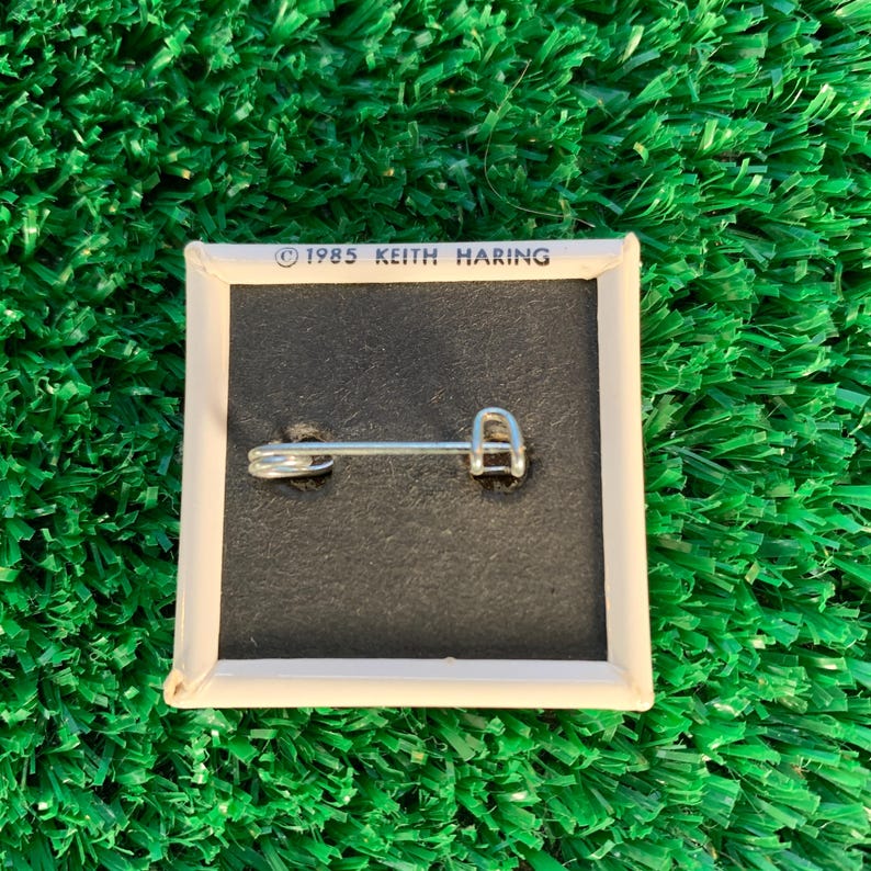 Puede incluir: Pin cuadrado con fondo negro y borde blanco. El pin tiene un cierre de imperdible plateado en la parte trasera. El texto "&copy; 1985 KEITH HARING" est&aacute; impreso en blanco en la parte superior. El pin est&aacute; sobre una superficie verde texturizada.