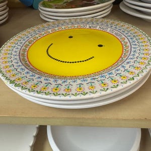May include: A stack of white ceramic plates featuring a yellow smiley face design. The plates have a floral border and the text "DO WHAT MAKES YOU HAPPY" around the edge. Other plates are visible in the background.