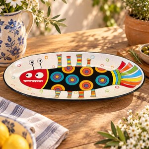 May include: An oval ceramic serving platter featuring a hand-painted creature with a red head, white eyes, and a black body decorated with colourful concentric circles. The creature has striped appendages and a multi-coloured tail on a white background.