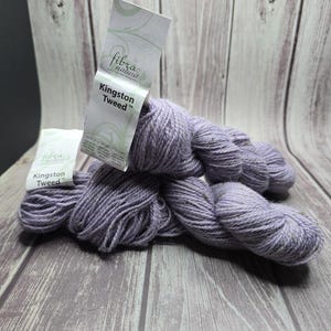 May include: Two skeins of lavender-colored Kingston Tweed yarn are displayed on a wooden surface. The yarn is a soft, textured material. The product tags are visible, with the brand name "fibra natura" and "Kingston Tweed" printed on them. The background is a light wooden surface.