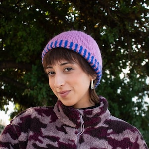 May include: A lilac knit beanie with blue stripes at the brim. The person is wearing a purple and burgundy camouflage fleece jacket. The background is a blurred view of green foliage.