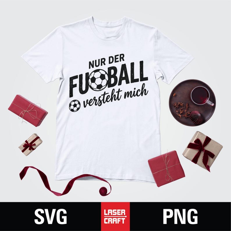 May include: White t-shirt with the text "NUR DER FUSSBALL versteht mich" in black, featuring a soccer ball graphic. The shirt is surrounded by wrapped gifts and a decorative plate with a drink.
