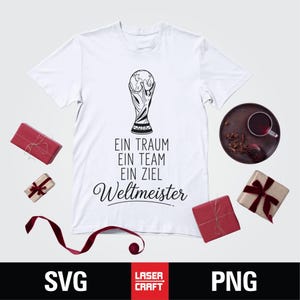 May include: White t-shirt with a black graphic of a trophy and the text "EIN TRAUM EIN TEAM EIN ZIEL Weltmeister". The shirt is surrounded by wrapped gifts and a decorative plate.
