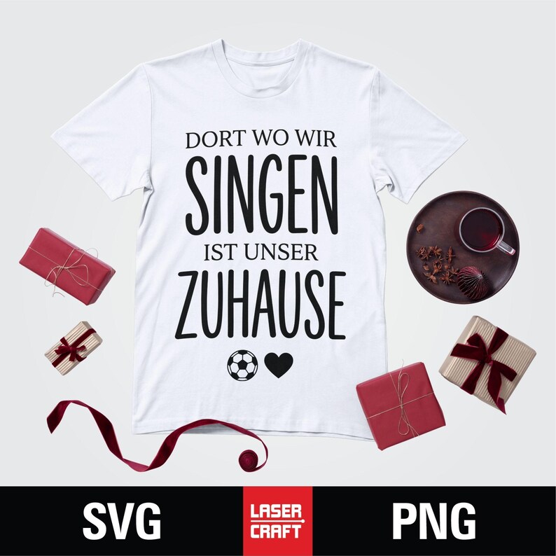 May include: White t-shirt with the German text "Dort wo wir singen ist unser Zuhause" (Where we sing is our home). The design includes a soccer ball and a heart. Several wrapped gifts and a drink are also visible.