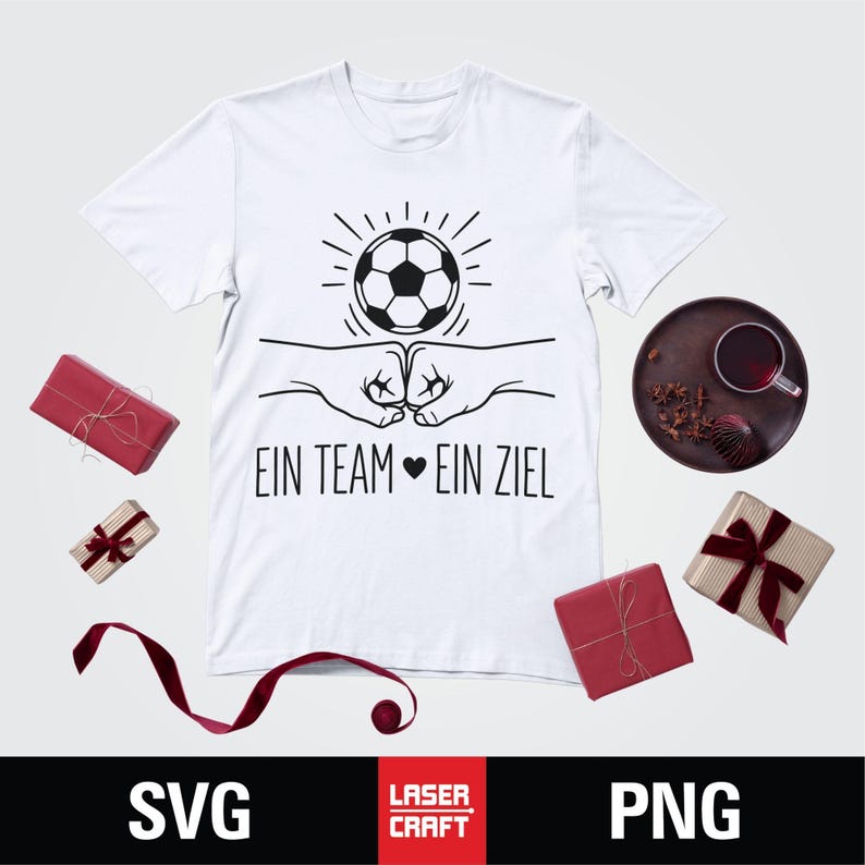 May include: White t-shirt with a black graphic of a soccer ball, two fists bumping, and the German text "EIN TEAM EIN ZIEL". The shirt is surrounded by wrapped gifts and a decorative plate.