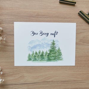 Puede incluir: Una tarjeta blanca con el texto "Der Berg ruft" en caligraf&iacute;a negra. La tarjeta presenta una ilustraci&oacute;n de acuarela de una cordillera con un bosque de &aacute;rboles verdes en primer plano.