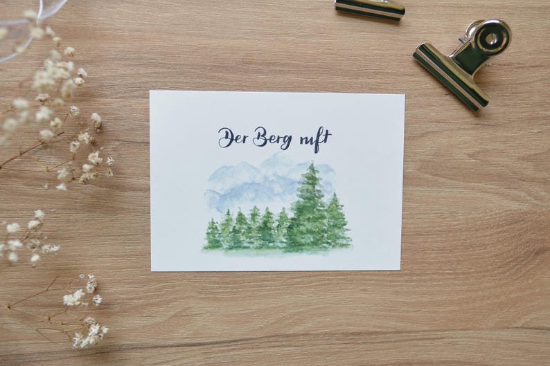 Puede incluir: Una tarjeta blanca con el texto "Der Berg ruft" en caligraf&iacute;a negra. La tarjeta presenta una ilustraci&oacute;n de acuarela de una cordillera con un bosque de &aacute;rboles verdes en primer plano.