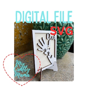 Puede incluir: Un marco de madera blanco con un diseño recortado de manos y la palabra "Papá". El marco está sobre un pequeño soporte de madera.