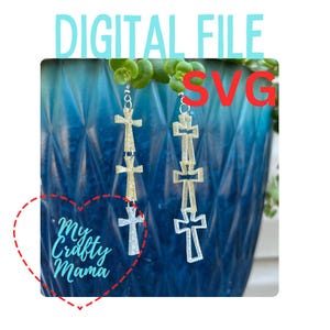 Op de afbeelding: Een paar zilveren en gouden kruis oorbellen met drie kruisen aan elke oorbel. De oorbellen hangen aan een plant in een blauwe pot. De tekst "My Crafty Mama" staat in een hart.
