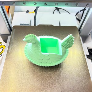 May include: A light green 3D-printed hen-shaped container with a square opening. The hen is on a gold-coloured textured surface, likely a 3D printer bed. The printer's components, including lights and wires, are visible in the background.