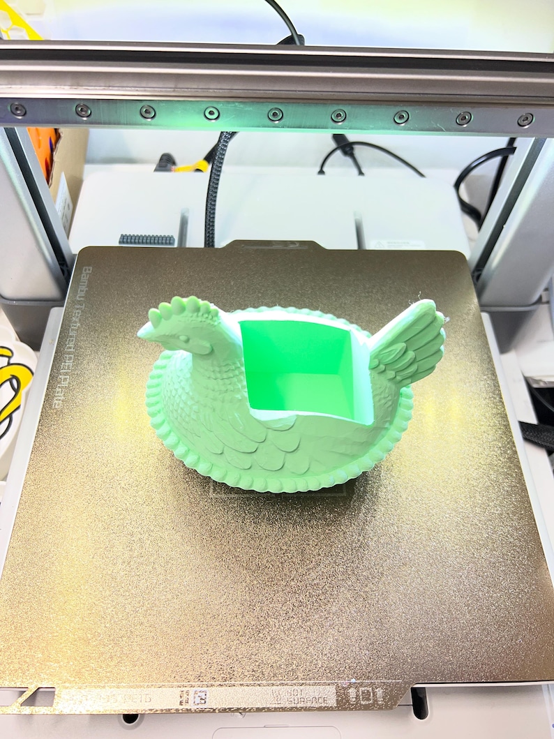 May include: A light green 3D-printed hen-shaped container with a square opening. The hen is on a gold-coloured textured surface, likely a 3D printer bed. The printer's components, including lights and wires, are visible in the background.