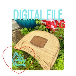 May include: A wooden cutout of a baseball cap with a brown patch on the front. The cap is sitting on a bed of green plants. The text "My Crafty Mama" is visible in the lower left corner of the image.